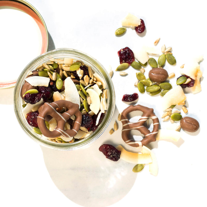 Top view of jar full of trail mix made up of milk chocolate pretzels and morsels along with seeds and coconut flakes