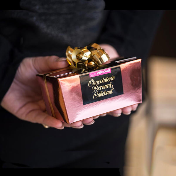 A Chocolaterie Bernard Callebaut® copper chocolate box with gold ribbon held behind someone's back