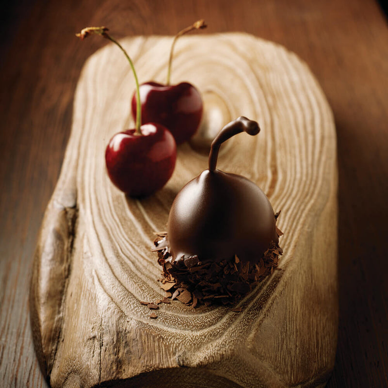One dark chocolate covered cherry and two fresh cherries sitting on a piece of wood
