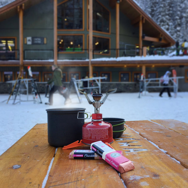 Camping burner with pot outside ski chalet with chocolate bars in forground